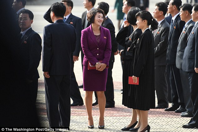 A young lady wearing a modern, brightly colored outfit and red clutch purse waits next to other women in traditional Korean dresses in Pyongyang
