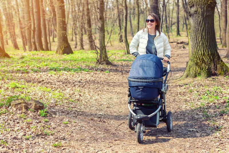Beautiful young adult woman walking with baby in stroller through forest or park on bright sunny day. Healthy lifestyle and stock image
