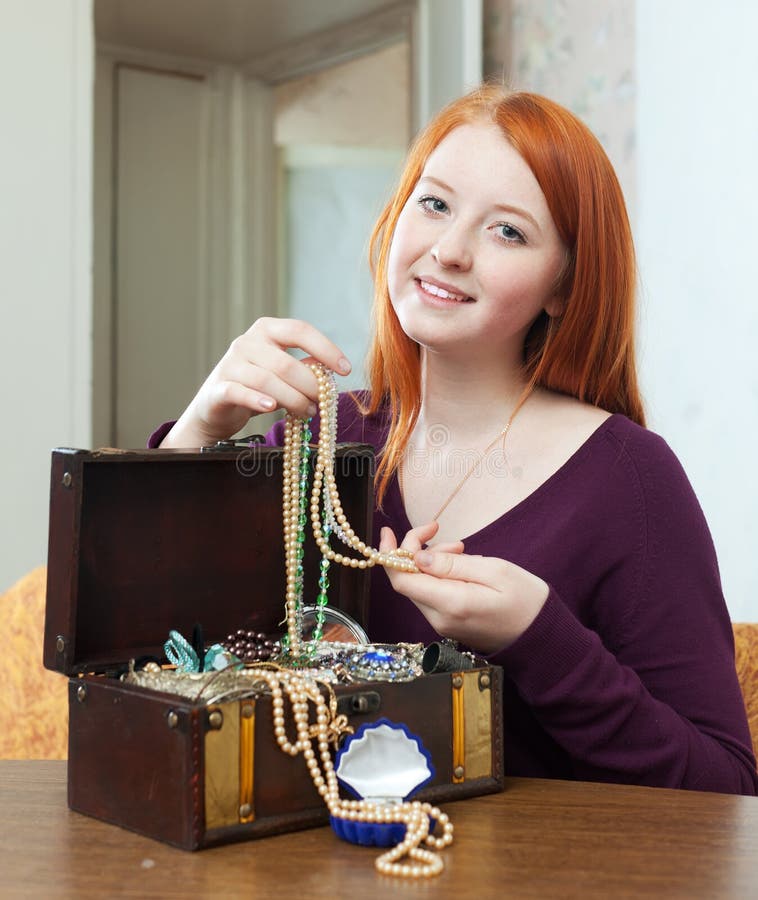 Teenager girl looks jewelry in treasure chest. Portrait of red-headed teenager girl looks jewelry in treasure chest at home interior stock photo